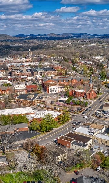 Aerial view of a small town with roads, buildings, trees, and hills in the background under a partly cloudy sky.
