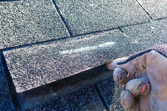 Close-up of a gloved hand lifting a roof shingle with a chalk line visible underneath on asphalt shingles.