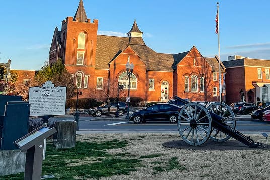 Historic brick building with a cannon in front, a Chickasaw Treaty Council marker, and cars parked nearby under a clear sky.