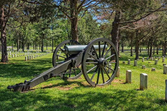 Historic cannon displayed among rows of graves and trees in a peaceful cemetery on a sunny day.