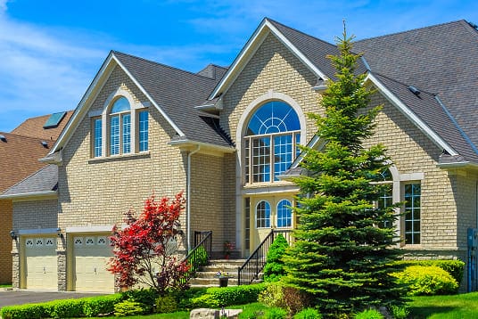 Large beige brick house with arched windows, a three-car garage, and a landscaped yard with trees and shrubs under a blue sky.