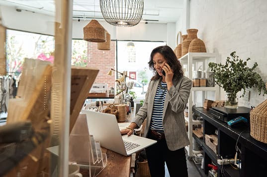Person standing at a wooden counter with a laptop in a bright, cozy store filled with wicker baskets and home decor items.