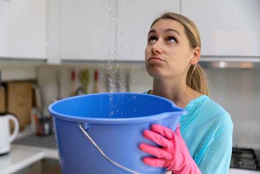 Person wearing pink gloves holding a blue bucket catching water leaking from above in a kitchen setting.