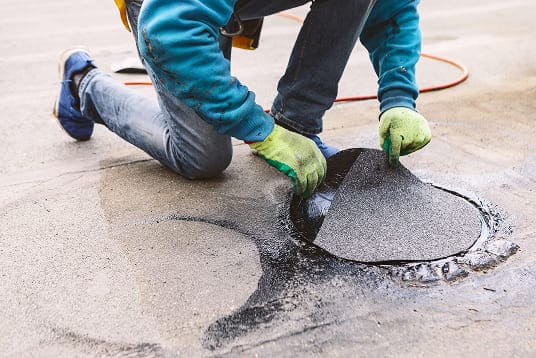 Roofer wearing gloves kneels while applying a circular patch of granular waterproofing material over wet sealant on a flat roof surface.
