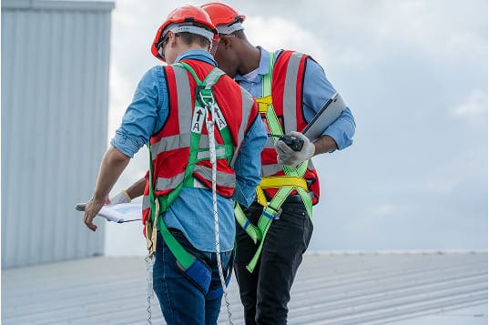 Two construction workers wearing hard hats, reflective vests, and bright green fall protection harnesses stand on a metal roof reviewing documents under a cloudy sky.